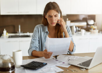 Candid shot of worried young European female dressed casually sitting at kitchen table, holding paper sheet and talking to house and utilities service representative about miscalculation in bills
