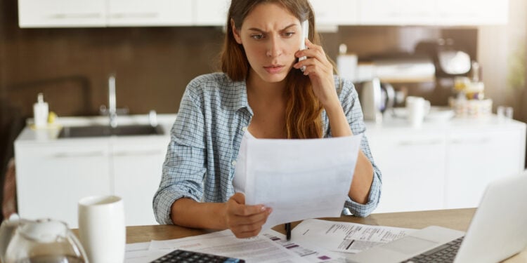 Candid shot of worried young European female dressed casually sitting at kitchen table, holding paper sheet and talking to house and utilities service representative about miscalculation in bills