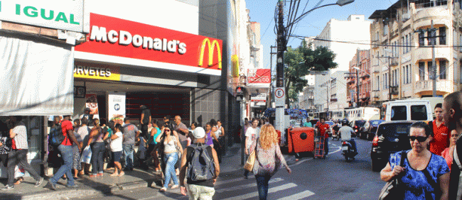 Clientes fazem fila na porta de lanchonete para comprar sorvete em meio ao tráfego intenso da via