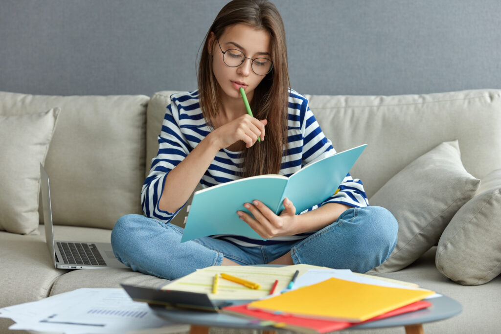 estudando o conceito online mulher jovem seria ocupada com um projeto freelance remoto senta se em um sofa confortavel faz anotacoes segura o livro usa o laptop em casa com internet sem fio