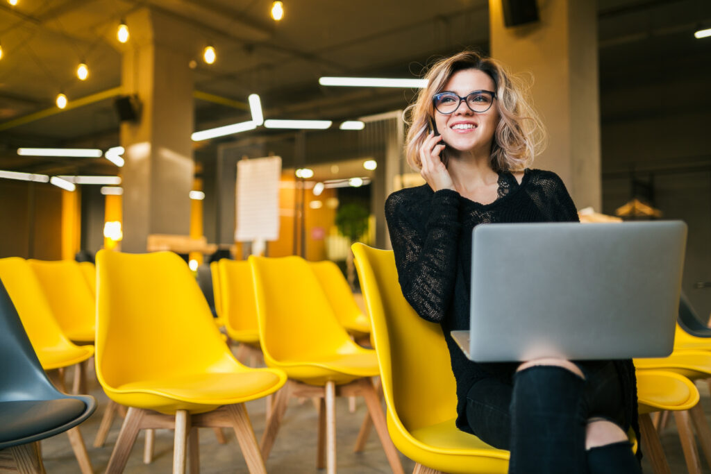 A romantização do “trabalhe de qualquer lugar”: o que ninguém fala sobre liberdade profissional 1 young attractive woman sitting lecture hall working laptop wearing glasses many yellow chairs student education online freelancer smiling talking smartphone looking forward startup