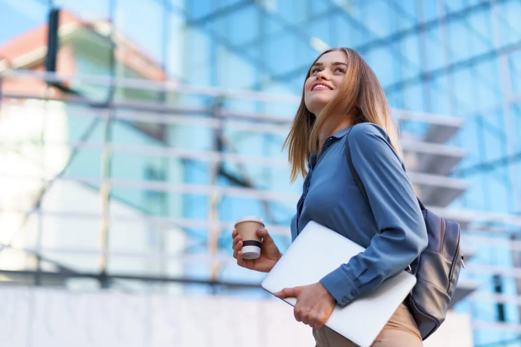 blonde young woman smiling portrait holding laptop coffee wearing blue gentle shirt modern building