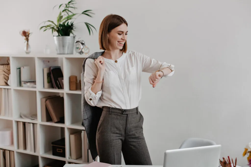 Como os hábitos moldam carreiras sem que percebamos 1 laughing business woman white blouse gray pants is looking wrist watch standing against office furniture