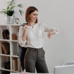 laughing business woman white blouse gray pants is looking wrist watch standing against office furniture